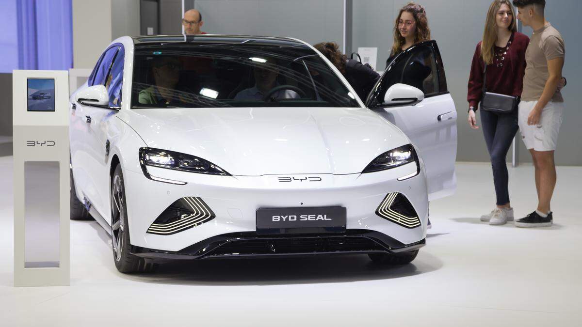 Vehículo eléctrico BYD Seal de color blanco exhibido en un salón de automóviles. El coche tiene un diseño moderno con faros delanteros estilizados y detalles luminosos en el parachoques. A la izquierda del vehículo hay un panel informativo de la marca BYD. Varias personas están observando y explorando el interior del coche, con una puerta abierta. La escena refleja un entorno de exhibición automotriz, probablemente en una feria o evento del sector.